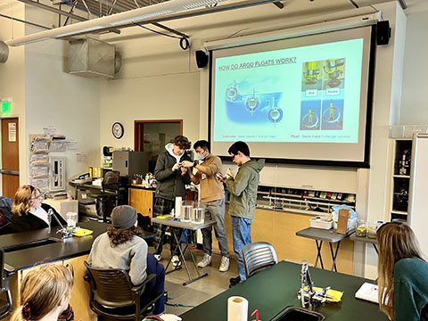 Three GOBOP members at the front of a classroom, demonstrating float buoyancy