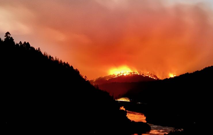 Glowing red fire and dense smoke rise above the silouette of tree-covered hills