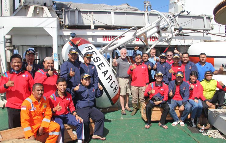 Group photo of 21 folks smiling and giving the thumbs up while on the deck of a ship in front of a mooring that says "research"