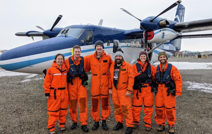 A group photo of  six scientists wearing orange full body flight suits, boots and some in beanies smiling in front of a blue and white NOAA plane with two propellers. 