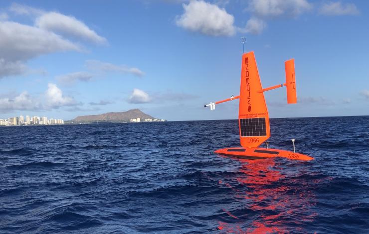 Orange drone with sail in open water in front of some bluffs and coastal city  in the distance