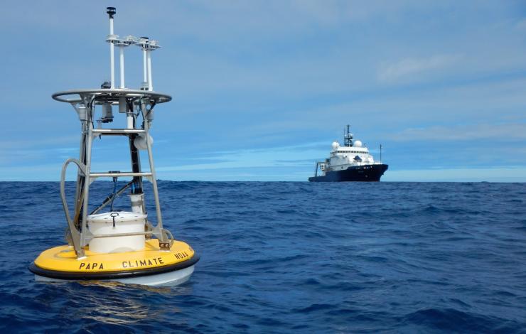 Yellow buoy floating on top of the water with a metal cage above with various instruments with a ship in the background