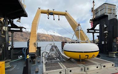 A yellow and white painted circular donut shaped mooring upside down on the back of a ship with the sea and landscape in the background