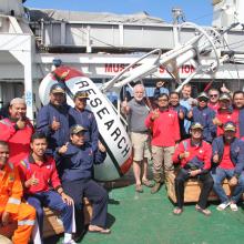 Group photo of 21 folks smiling and giving the thumbs up while on the deck of a ship in front of a mooring that says "research"