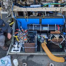 Dave squatting to the left of a remotely operated vehicle with a blue hat and mask on. Variety of tubes, bottles and sensors are shown as part of the ROV
