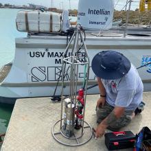 A person with a blue rimmed hat with gray shirt and camouflage pattern shorts kneeling in front of a metal cage hosting 3 silver instruments with a toolbox nearby in front of the uncrewed surface vessel with water behind. 