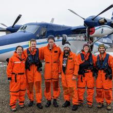 A group photo of  six scientists wearing orange full body flight suits, boots and some in beanies smiling in front of a blue and white NOAA plane with two propellers. 