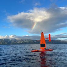 Orange drone in the middle of blue water with clouds above and a rainbow in the background