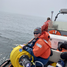 Grey skys with Angie in a beanie and orange life jacket sitting next to a yellow buoy on a rigid inflatable boat