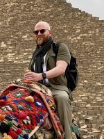 Doug in a black shirt and khaki pants sitting on top of a camel in front of the pyramids 