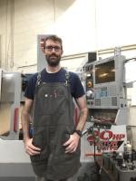 Sean standing in front of machine with grey work apron and glasses on 