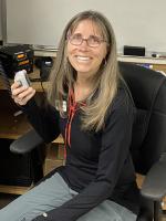 Diane sitting on a chair holding in her right hand a HAM radio with a whiteboard and desk behind her