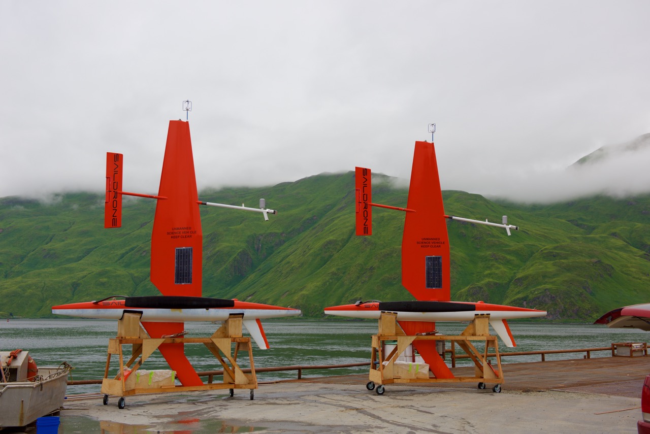 Two saildrones on wooden racks sit near the water, vibrant green mountains and dense clouds in the distance.