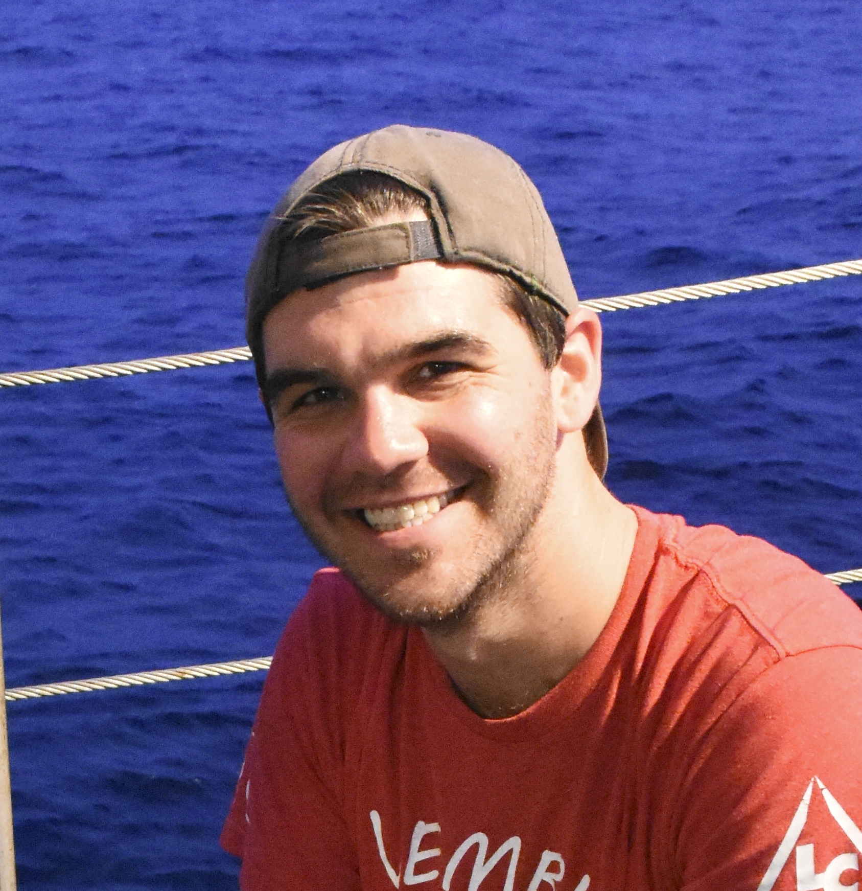 Jonathan smiling in red shirt and backwards hat in front of water